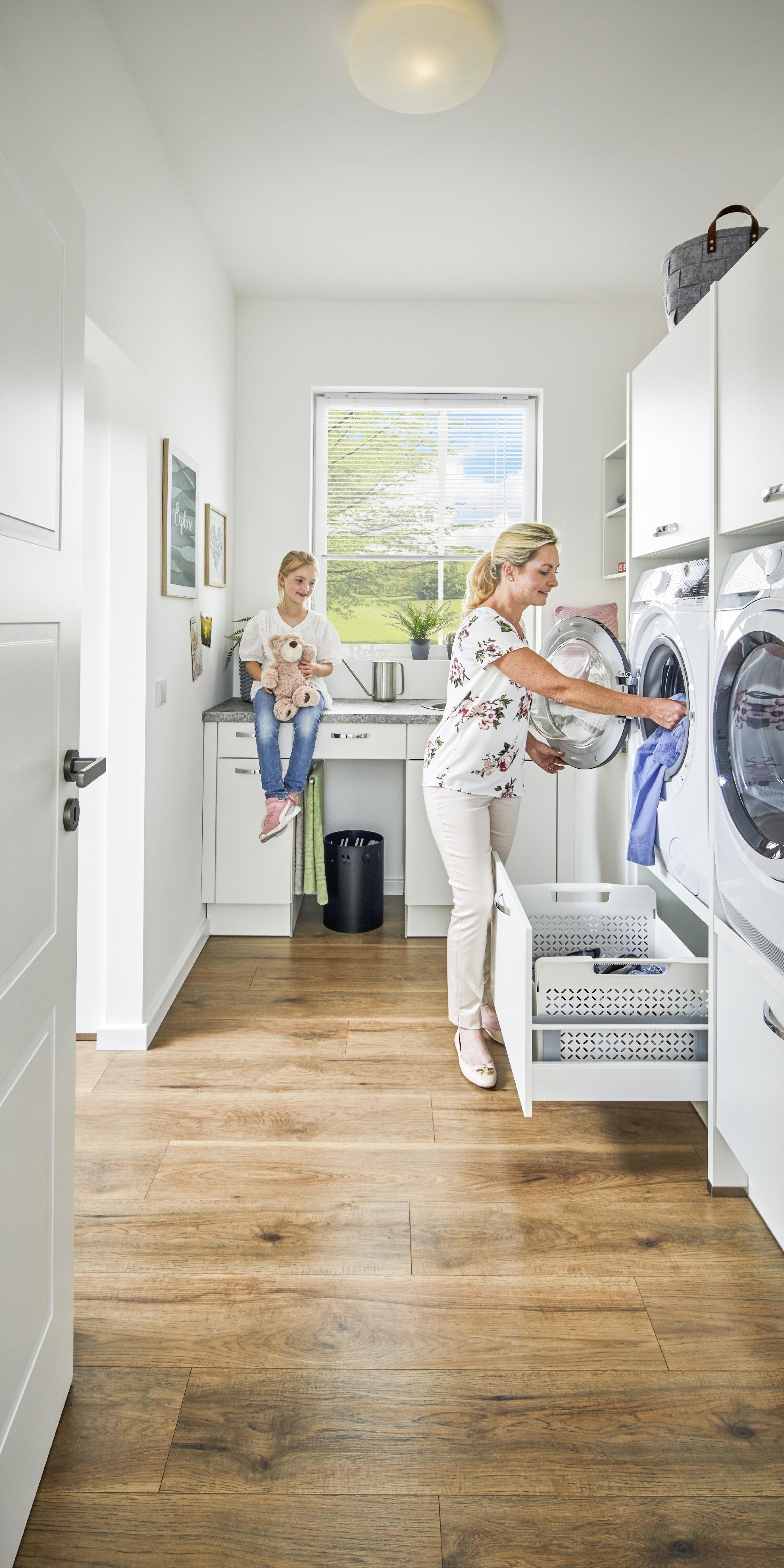 schmaler Hauswirtschaftsraum ergonomisch erhöht eingebaute Waschmaschine und Trockner Schmutzwäsche-Schublade geöffnet Mutter gibt daraus Wäsche in Waschmaschine Kind sitzt daneben auf Spül-Schrank