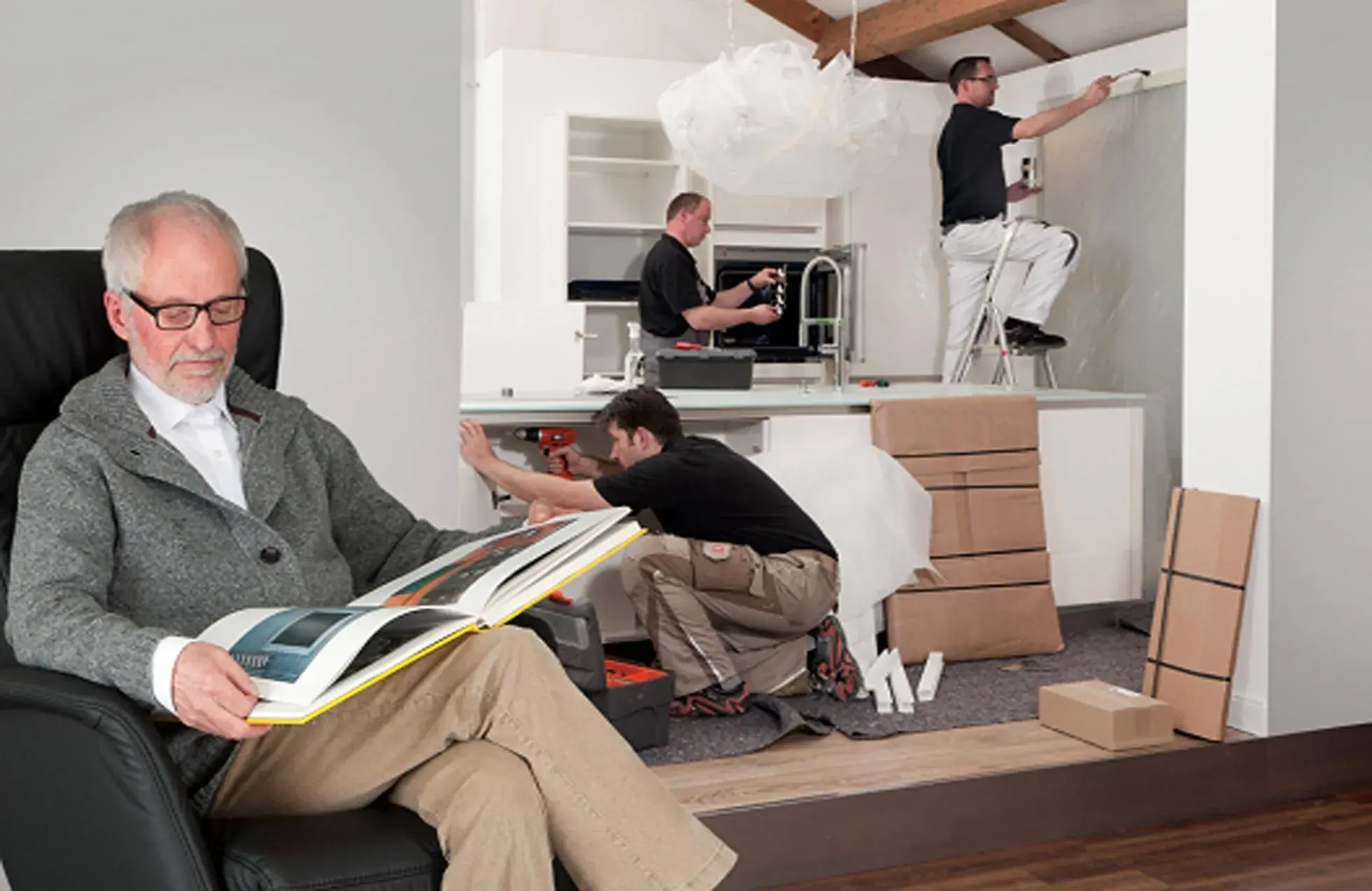 Mann mit Buch im Lesesessel vor drei Monteuren bei der Küchenmontage im Hintergrund